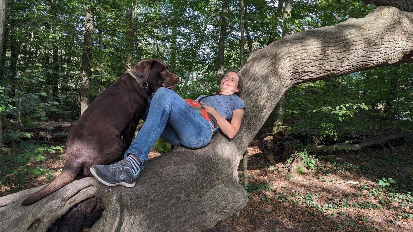 Tanja entspannt mit ihrem Hund auf einem liegenden Baumstamm im Wald